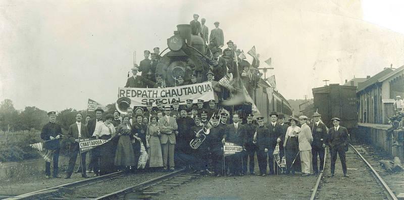 large group of people from the 1800s gathered around the top and around a train at a train station. The people have musical instruments and signs