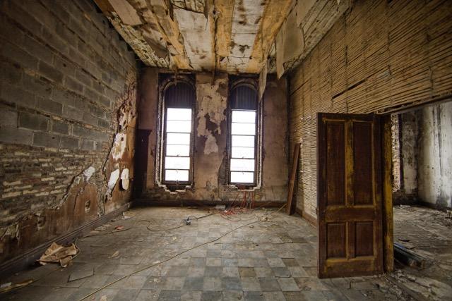 Dilapidated room in Antoinette Hall. Two long vertical windows at front of room with a door leading to a hallway