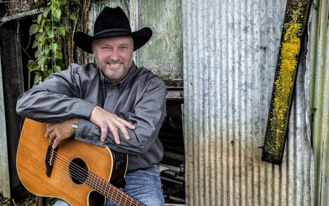 Jeff Carson sitting with his arms resting on a guitar in front of an old barn